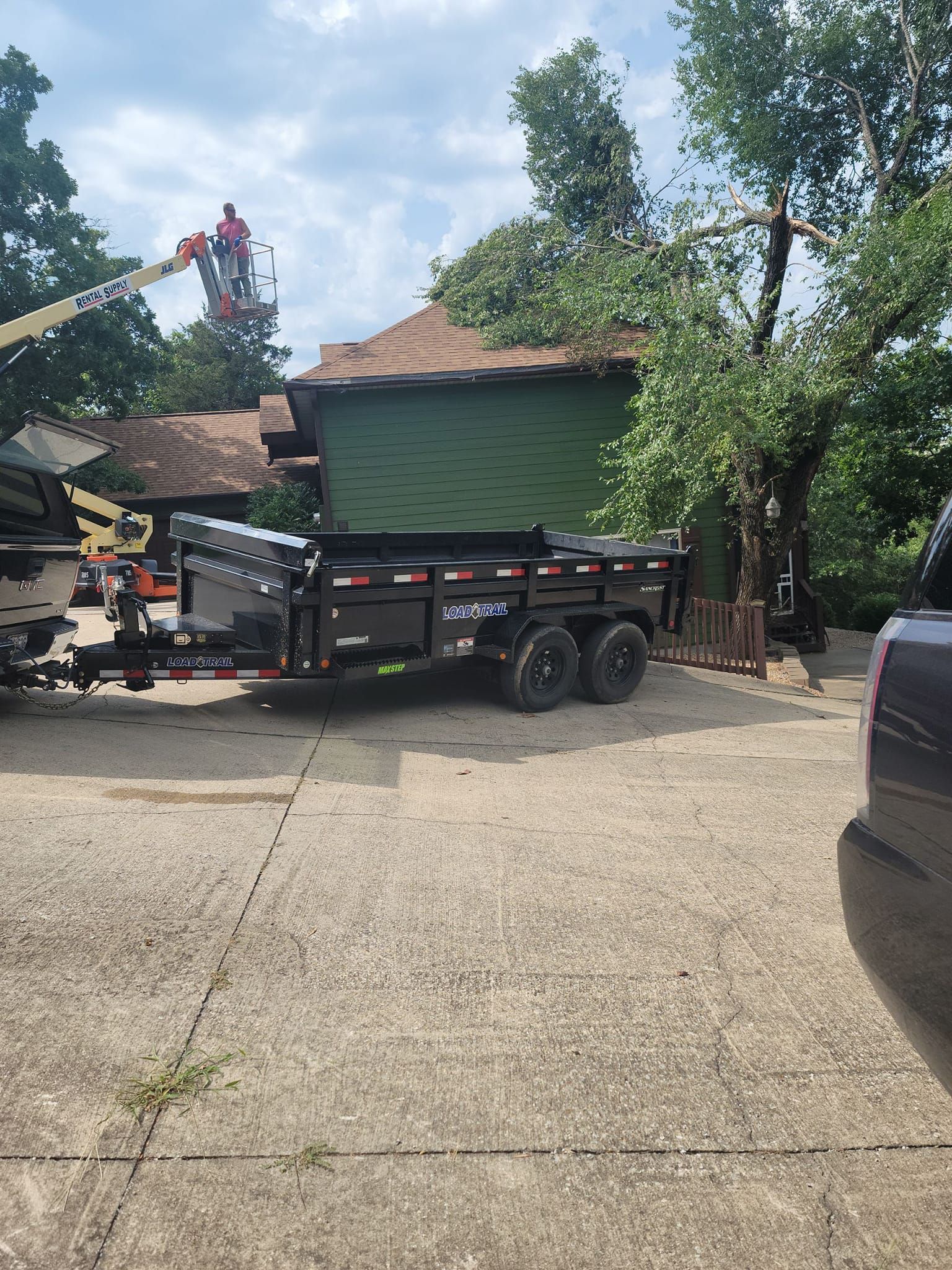 Black trailer in driveway near green house as a worker trims a tree from a lift.