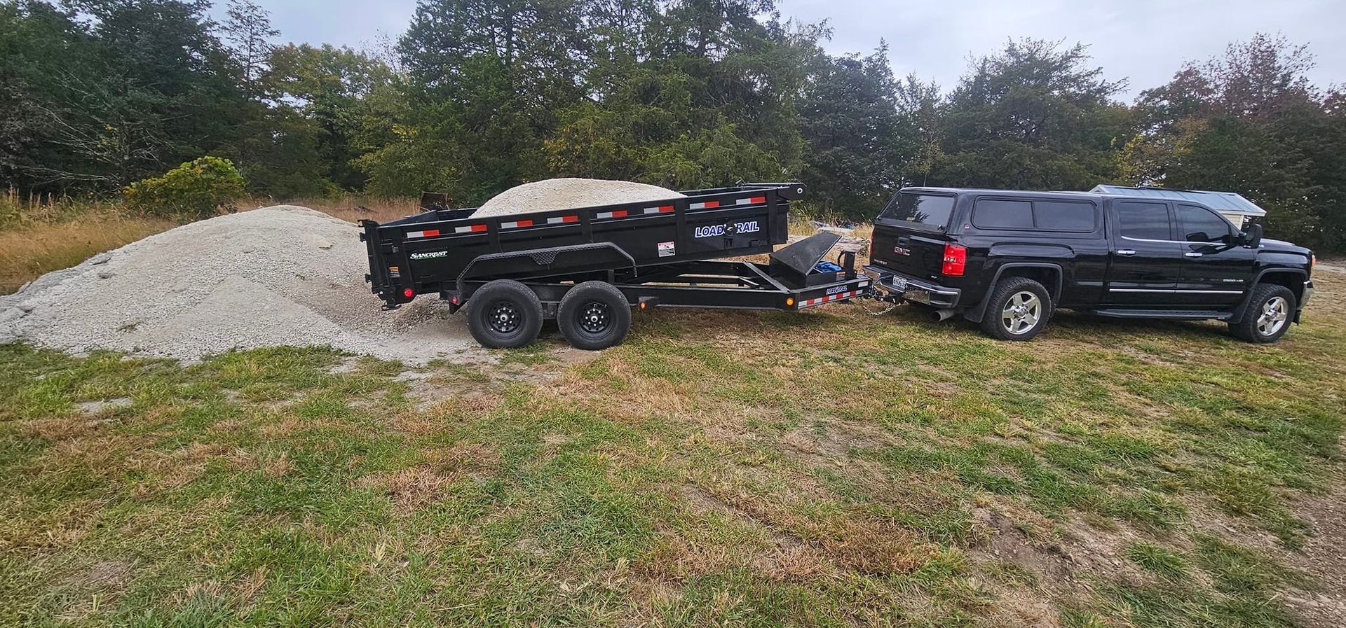 Black truck towing a trailer loaded with gravel on a grassy field, near a pile of gravel.