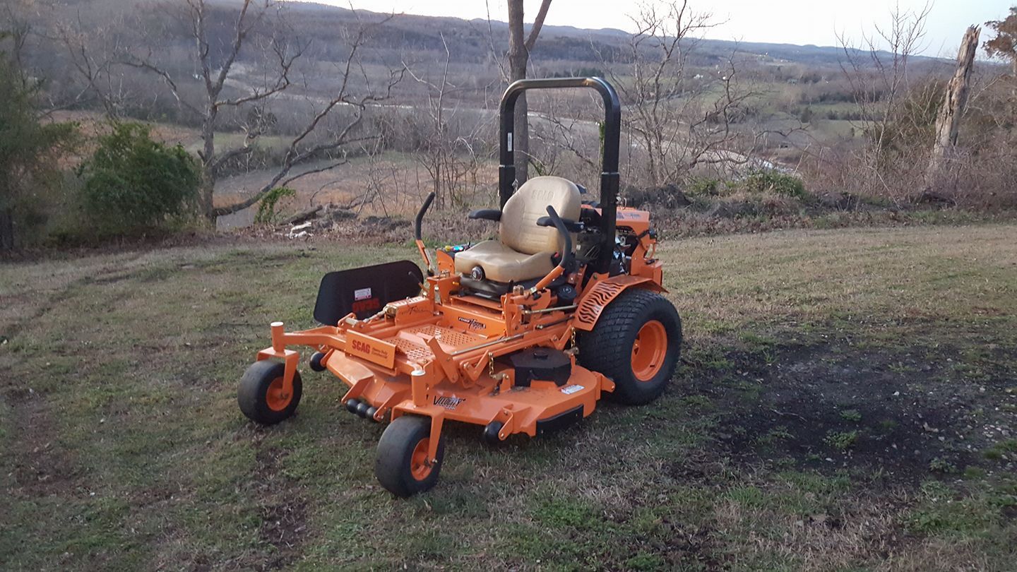Orange and black zero-turn mower on grass with a background of trees and a cloudy sky.