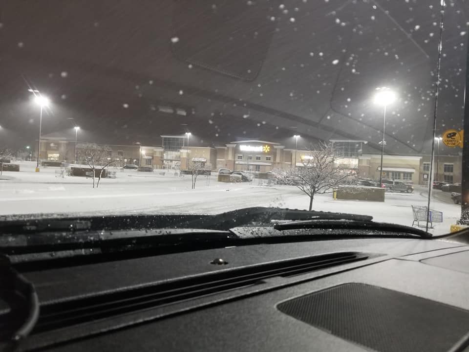 Snowy scene through a car windshield, streetlights illuminate a shopping center.