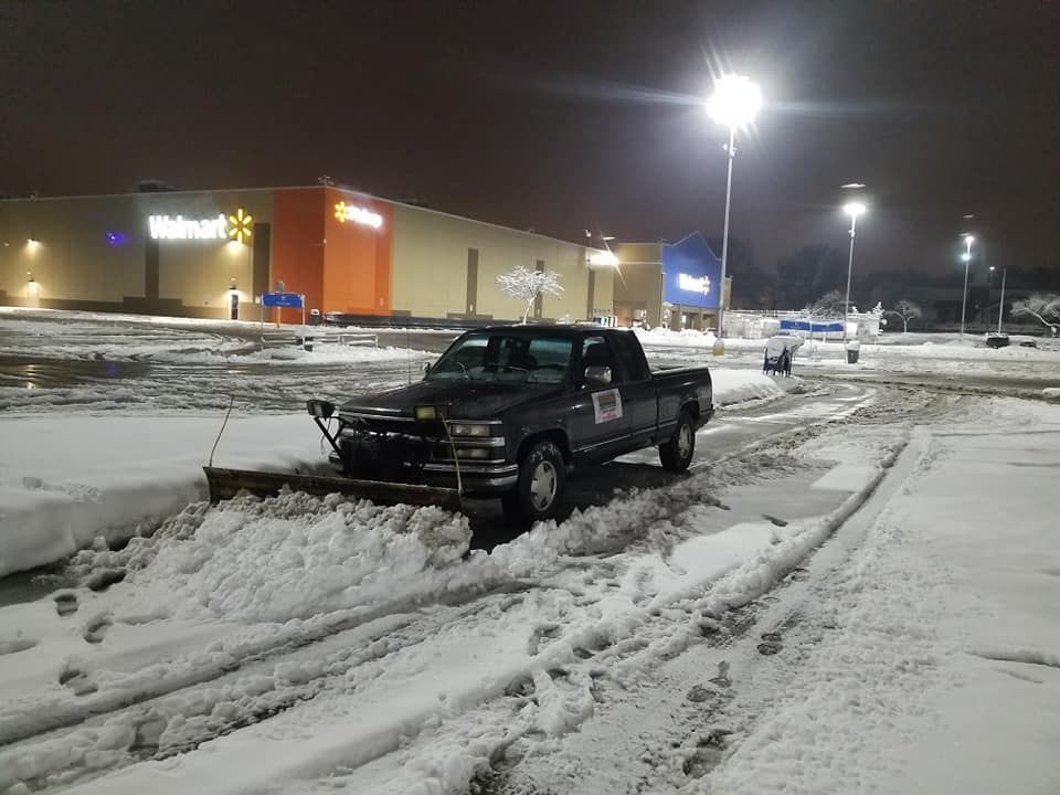 Snowplow truck clearing snow from a Walmart parking lot at night.