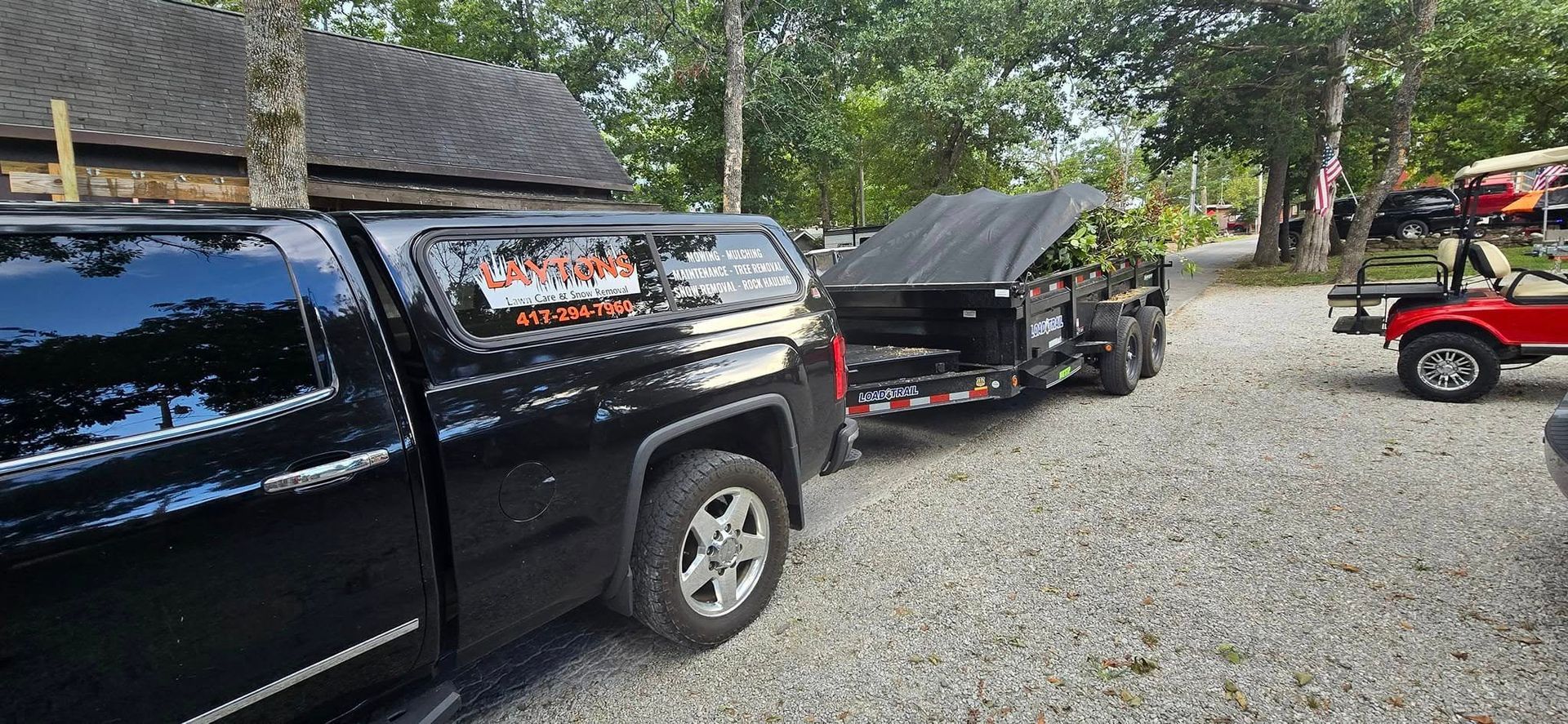 Black truck with a trailer, carrying debris, parked on gravel. A red golf cart is visible.