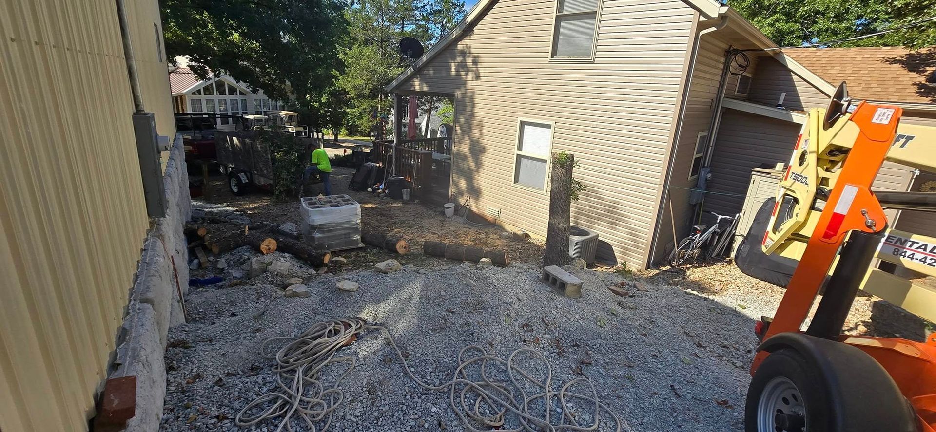 Construction site: tree debris and equipment next to a building with gravel and wooden logs.
