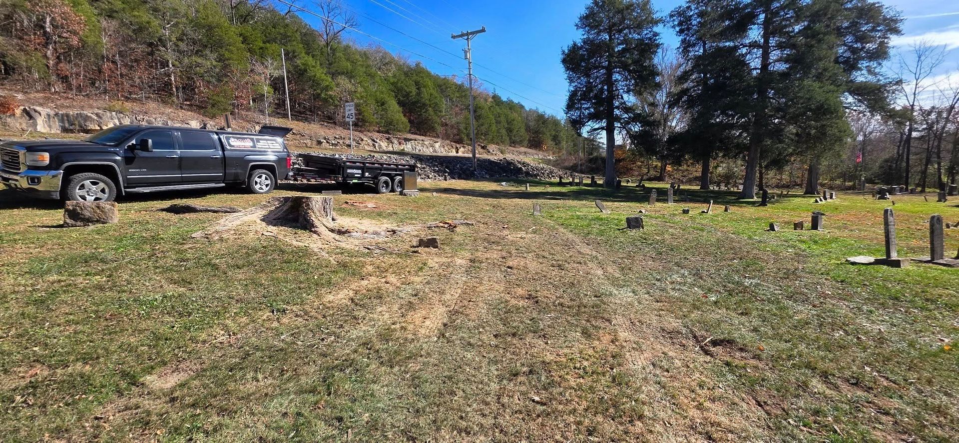 A black truck and trailer parked in a cemetery with a grassy foreground and a hill in the background.
