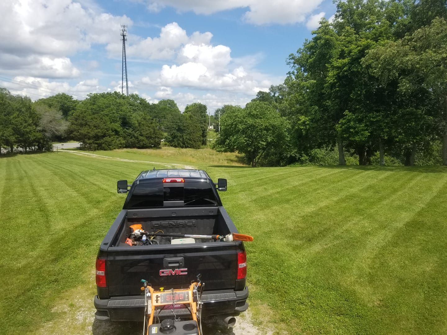 Black pickup truck with lawn equipment in the bed, mowing freshly striped green grass, trees in the background, blue sky.