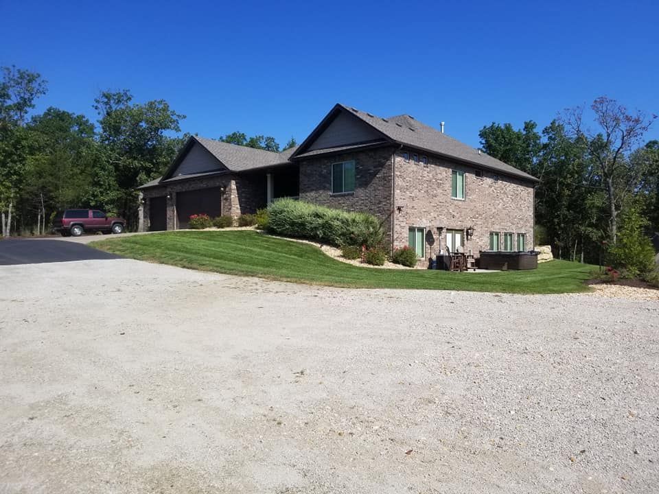 Two-story brick house with attached garage, gravel driveway, and green lawn under a blue sky.