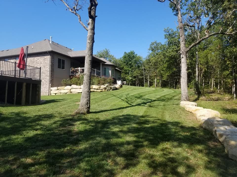 Lawn with a brick house and a deck. Trees flank the yard with a rock retaining wall.