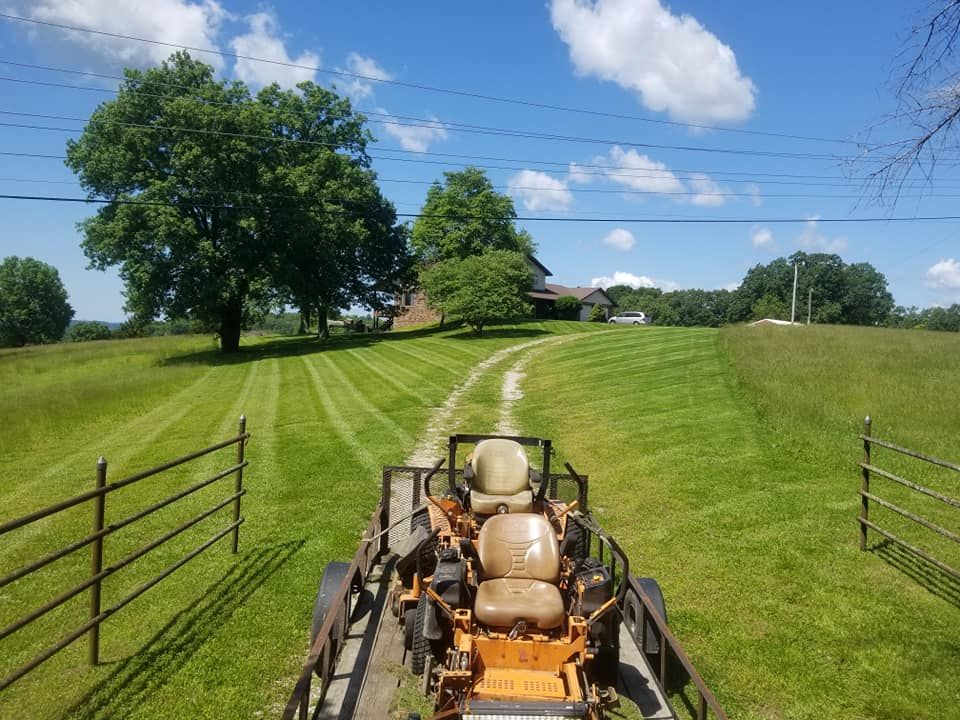 Lawnmower on trailer, following a grass path toward a house, on a sunny day.