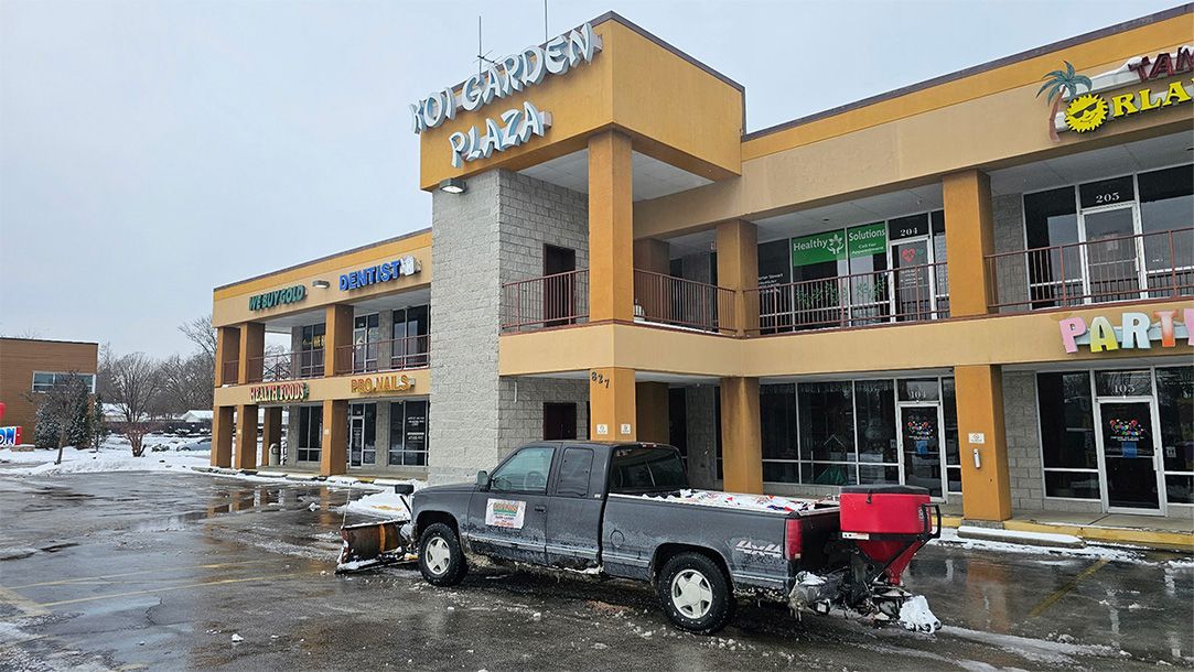 Truck plowing snow in front of a shopping plaza with businesses. Beige building, grey sky, winter scene.