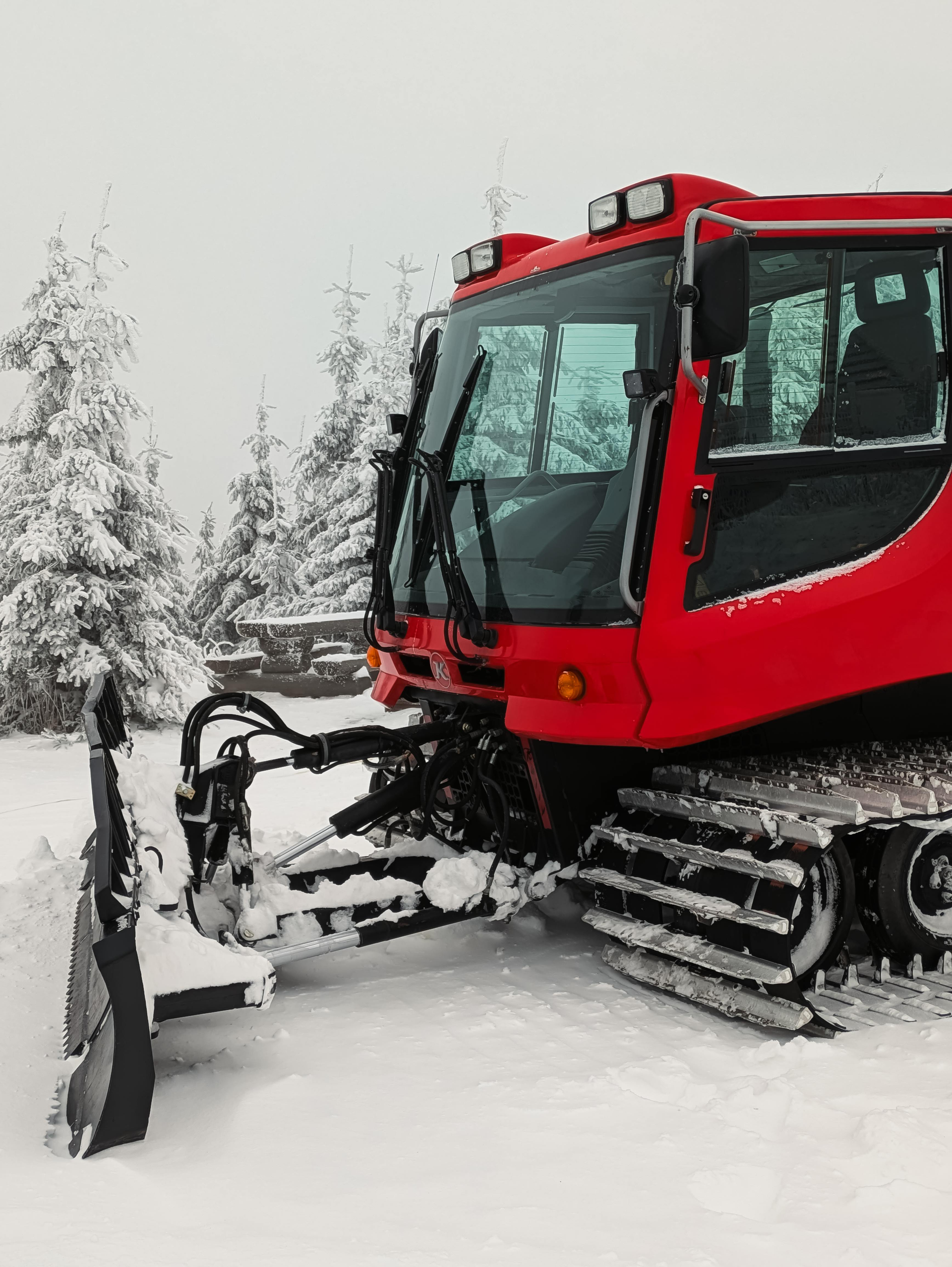 Red snow groomer on snow, clearing a path near evergreen trees.