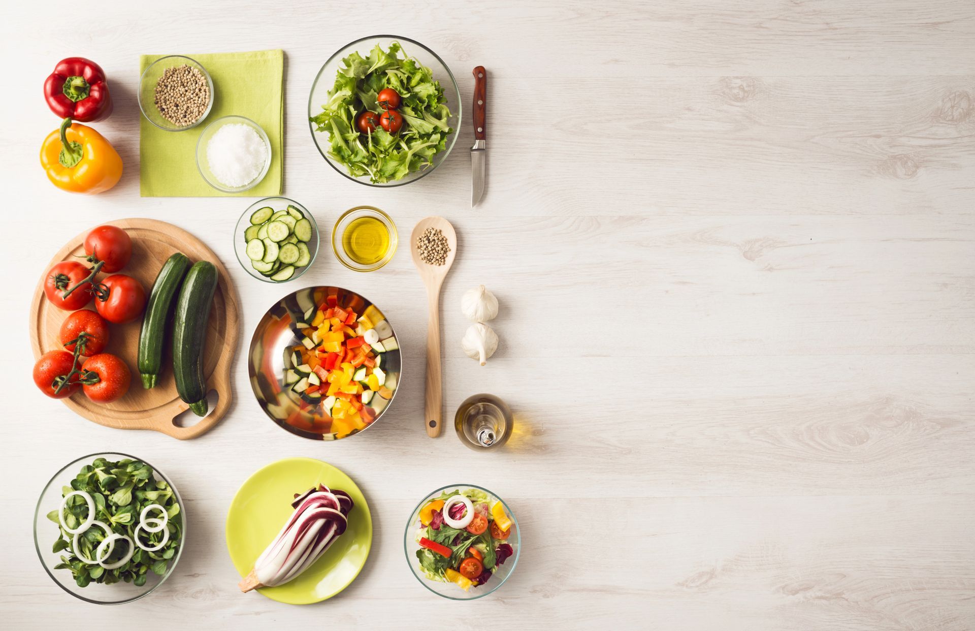 Vista dall'alto degli ingredienti freschi per un'insalata: verdure, olio ed erbe aromatiche disposti su un tavolo bianco.