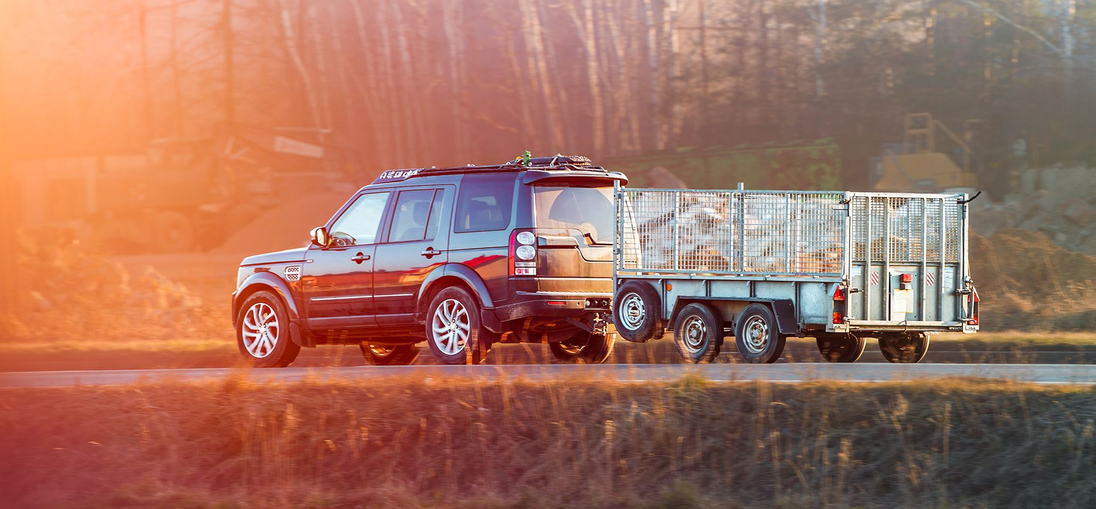 Dark SUV towing a trailer loaded with what appears to be wood, driving on a road in the countryside.