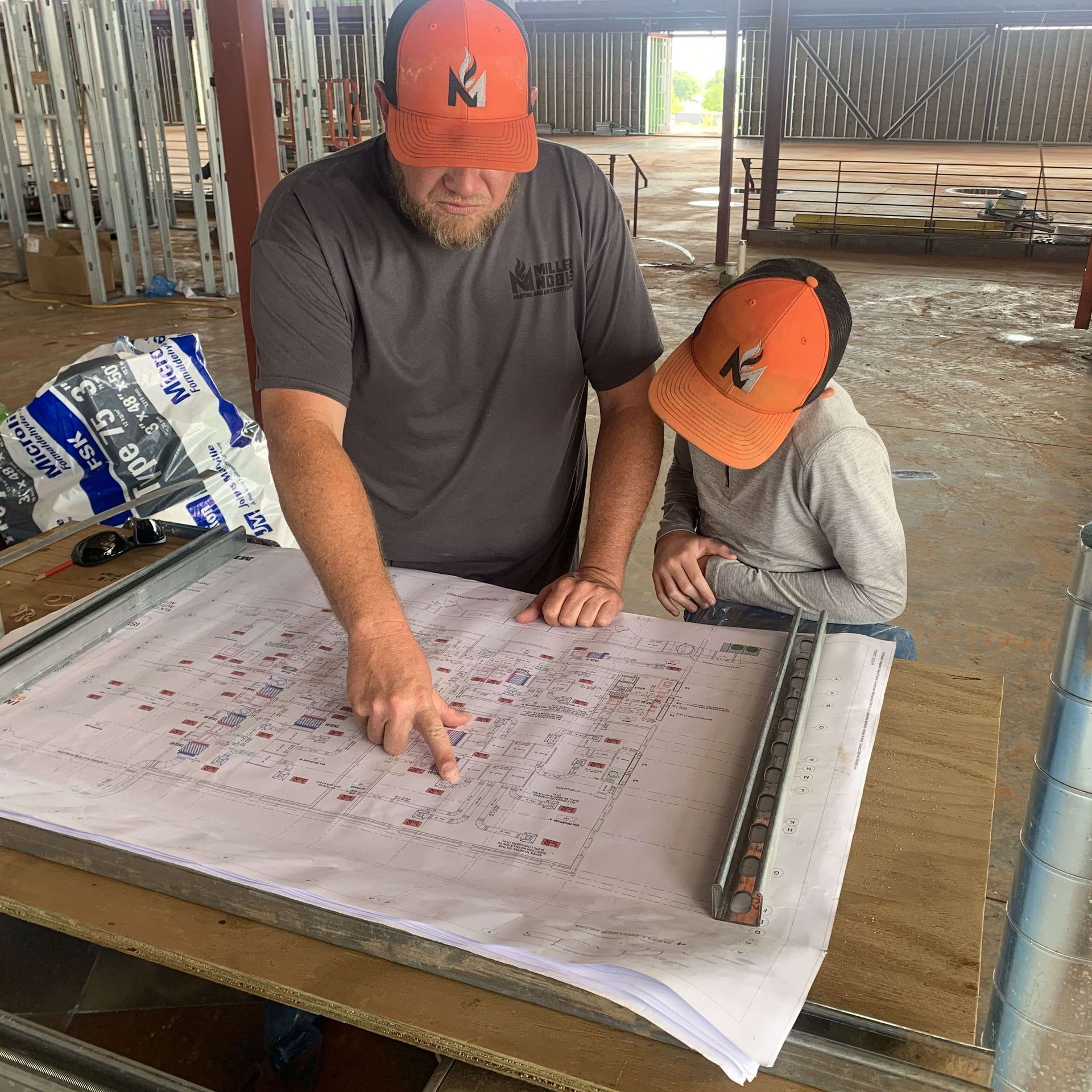 Two workers in orange hard hats review a large construction plan on a table inside a building under construction