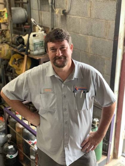 Man in a light gray work shirt standing in a workshop, smiling with hand on hip.