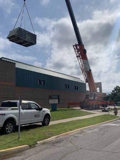 Crane lifting a large rooftop unit onto a brick building beside a parked white pickup truck.
