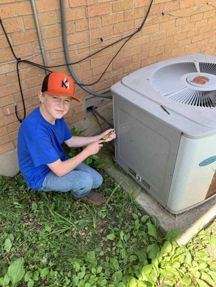 Boy in orange cap kneels by outdoor air conditioner beside a brick wall.