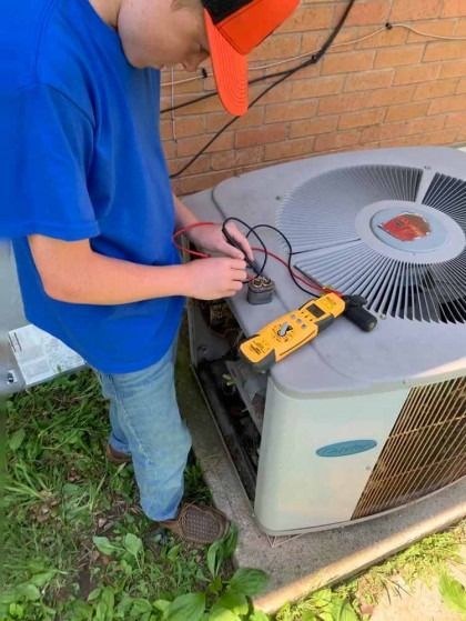 Technician testing an outdoor air conditioner with a yellow multimeter by a brick wall
