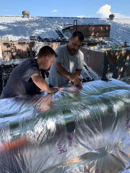 Two workers wrapping a large object in shiny silver insulation on a rooftop.