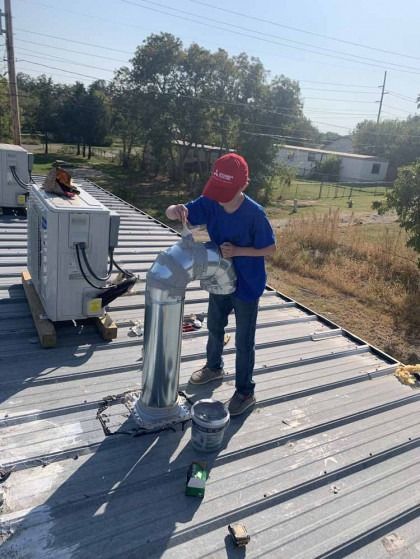 Worker in blue shirt on a rooftop installing a silver vent pipe near HVAC equipment.
