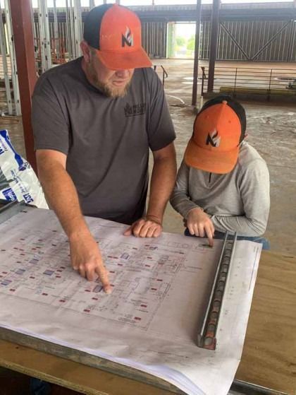 Two people in orange caps review a large printed sheet on a table, pointing at details.