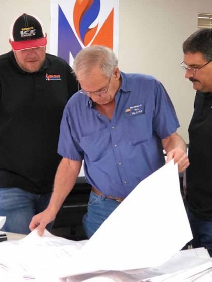 Three men in blue shirts examine large white plans at a table.