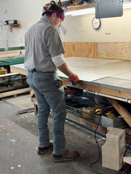 Person operating a table saw in a workshop, cutting wood beside stacked boards and cinder blocks.