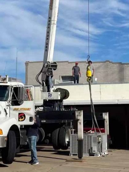 Crane lifting two workers on a platform beside a white truck at a construction site