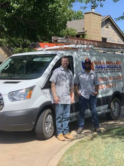 Two workers stand beside a white service van with ladders parked in a driveway under trees.