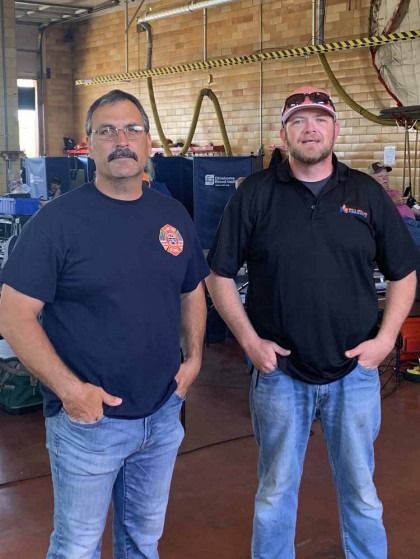 Two men in navy shirts and jeans standing in a workshop with brick walls and hanging tools.