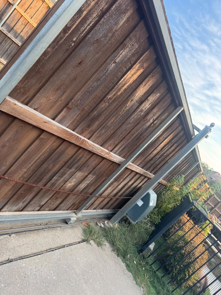 Tilted view of a wooden fence beside a sidewalk, with a trash bin and black gate under a cloudy sky
