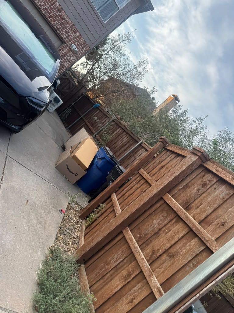 A tilted view of a driveway beside a wooden fence, with a parked car, a blue bin, and cardboard boxes.