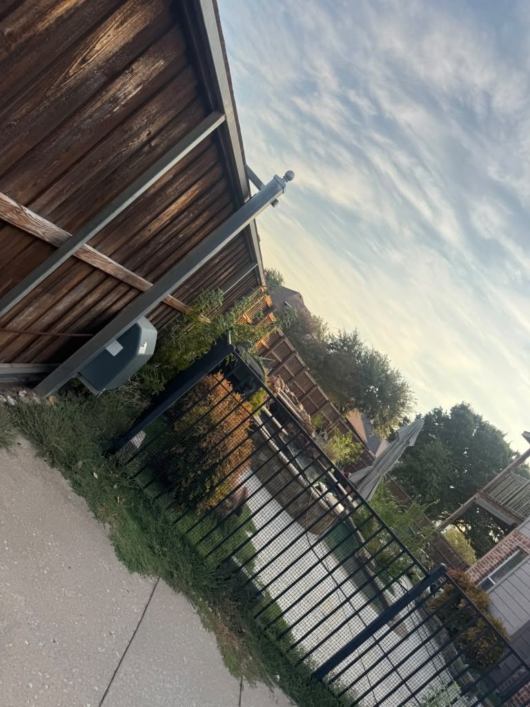 Tilted view of a wooden fence, black gate, and sidewalk beside a residential street under a cloudy sky