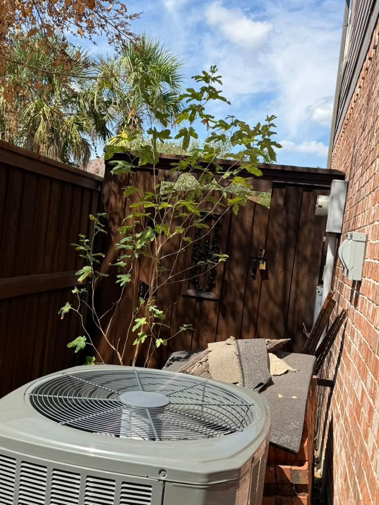 Backyard corner with an air conditioner unit, wooden fence, potted tree, and scattered boards in sunlight