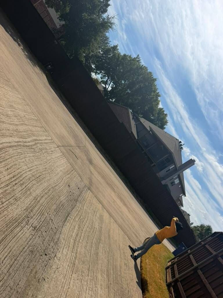 Person in yellow pants walking across a large paved lot beside a dark fence under a blue sky.