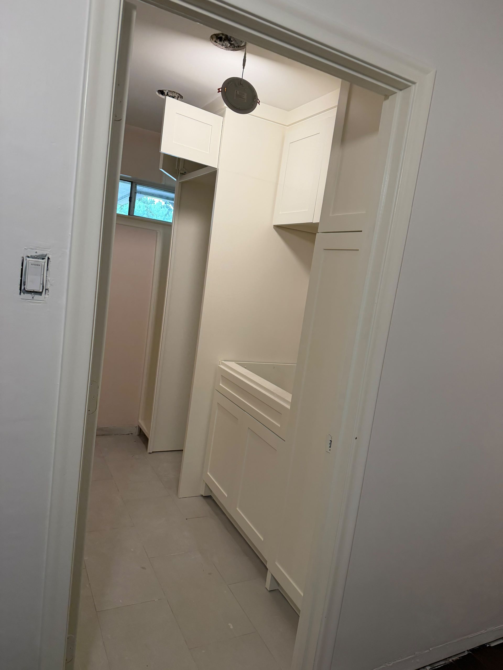 Narrow laundry or utility closet with white cabinets, radiator, and a window at the end of a hallway
