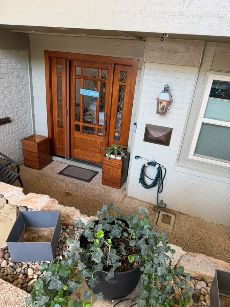 Front porch with wooden double doors, potted plants, a wall lamp, and a coiled hose by the entrance.