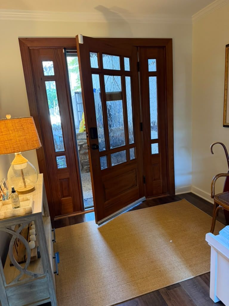 Wooden front door open to a bright entryway with a rug, side table, lamp, and chair