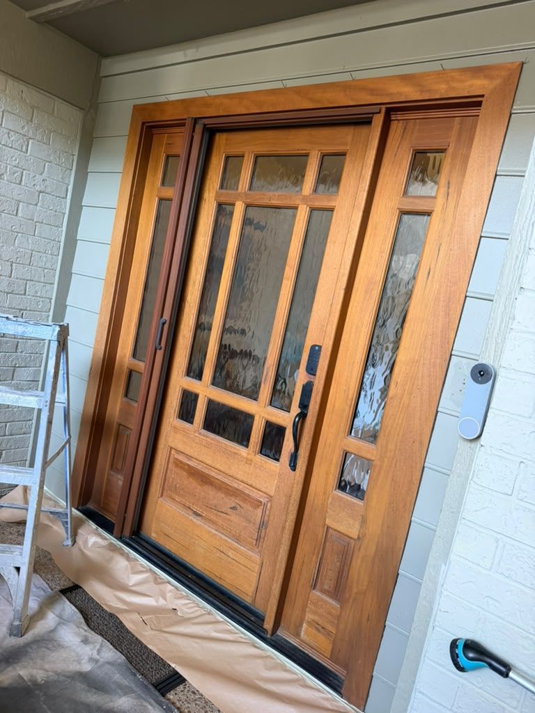 Wooden front door with glass panels and black handles, set in a covered entryway with protective floor covering