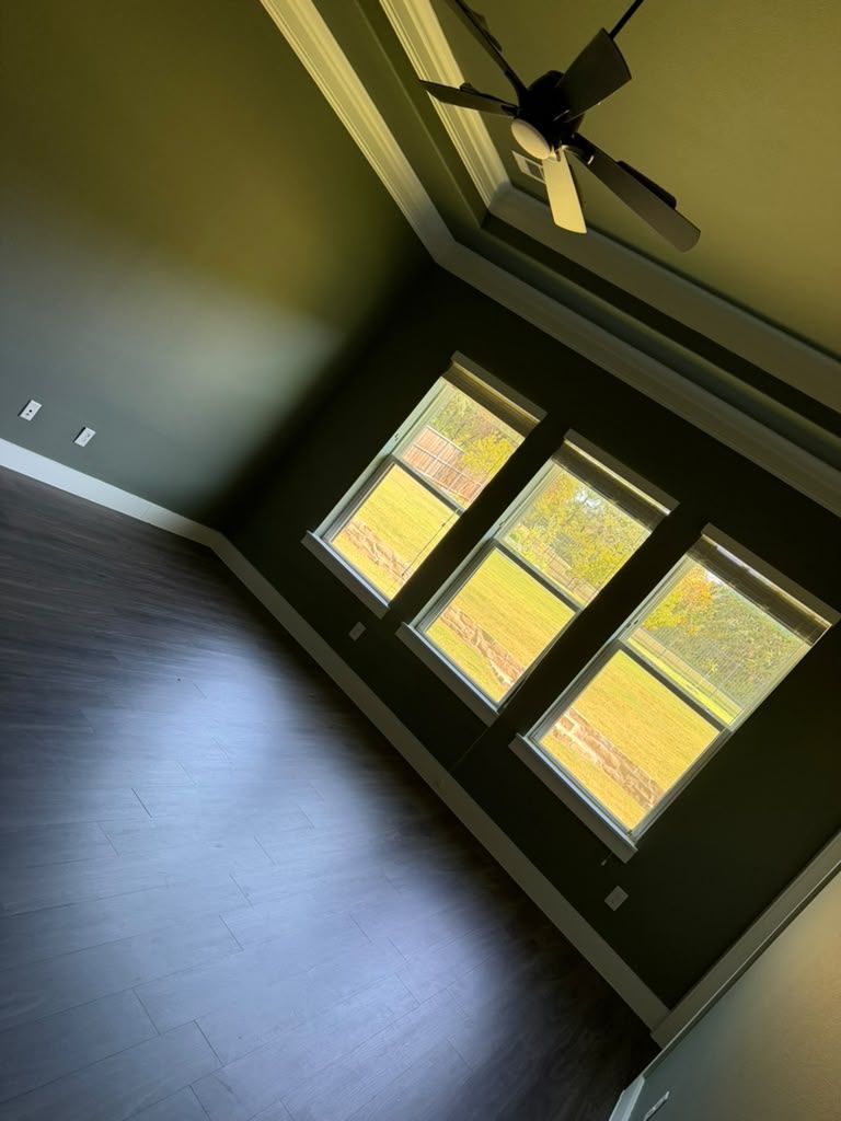 Empty room with dark wood floors, green walls, ceiling fan, and sunlight through large windows