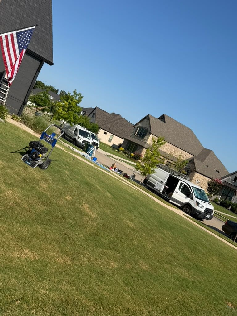 Suburban street with parked vehicles, a lawn mower on grass, and an American flag by a house under a blue sky