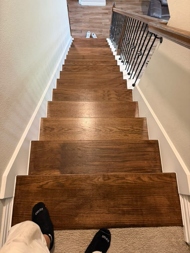 Looking down a wooden staircase with black railing, seen from the top with feet visible in foreground.