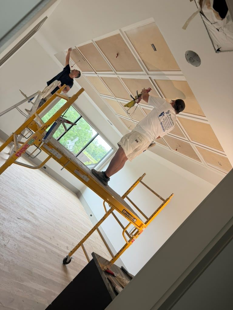 Two workers on yellow scaffolding painting a white ceiling in a room with large windows.