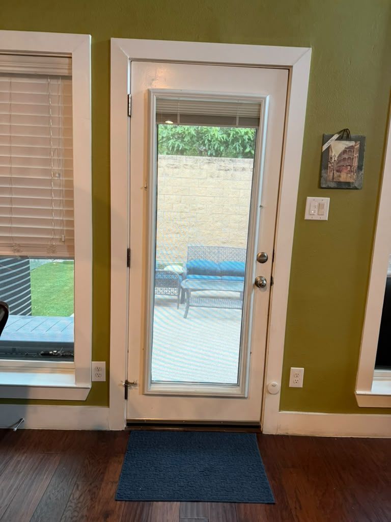 Indoor view of a glass patio door between windows, showing a deck and outdoor chairs beyond.