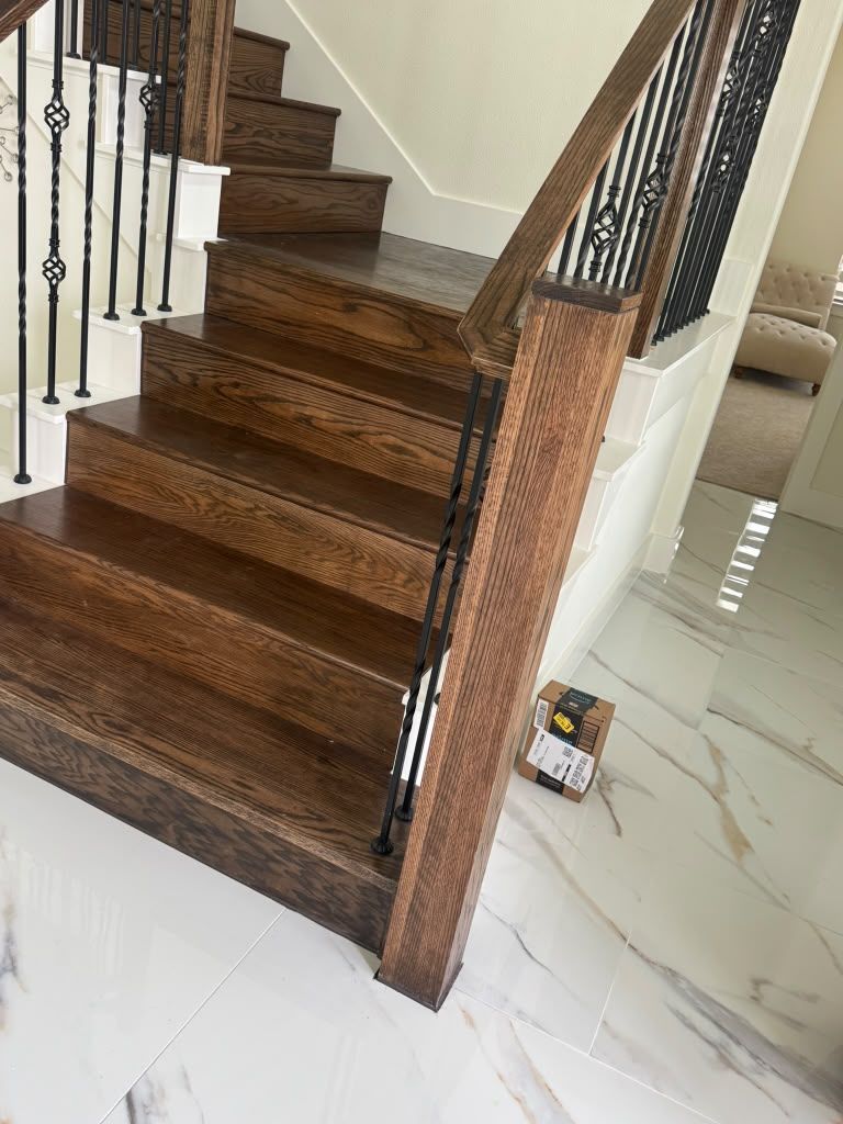Wooden staircase with black iron railing beside a white marble floor, in a bright interior.