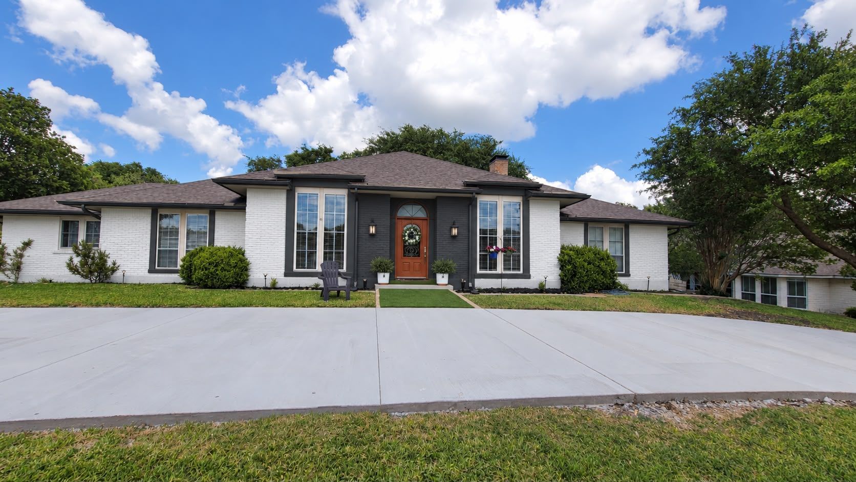 Single-story white house with dark shutters, front walkway, and a wide driveway under a partly cloudy sky