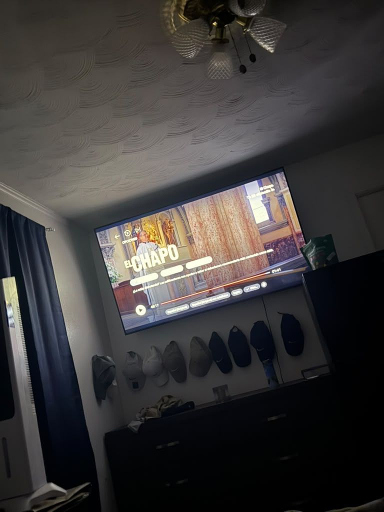 Bedroom with a TV showing a movie, ceiling fan overhead, and clothes hanging on the wall.