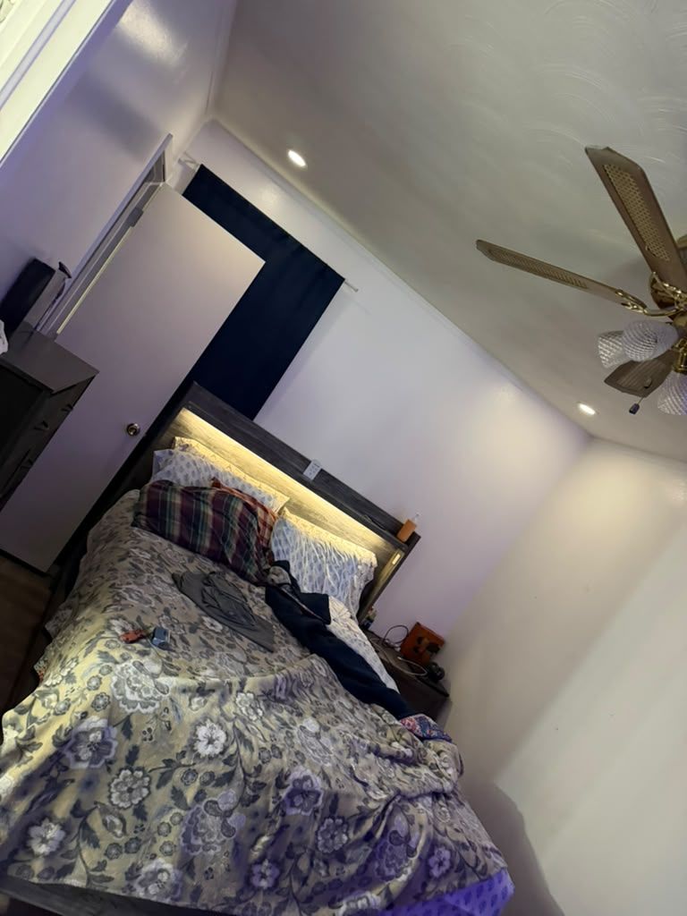 Bedroom with an unmade bed, gray patterned comforter, lit headboard, white walls, and a ceiling fan