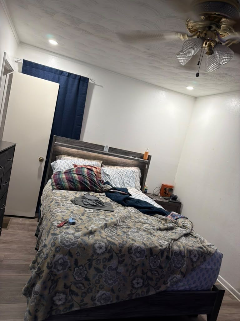 Bedroom with unmade bed, blue curtains, white walls, and a ceiling fan overhead