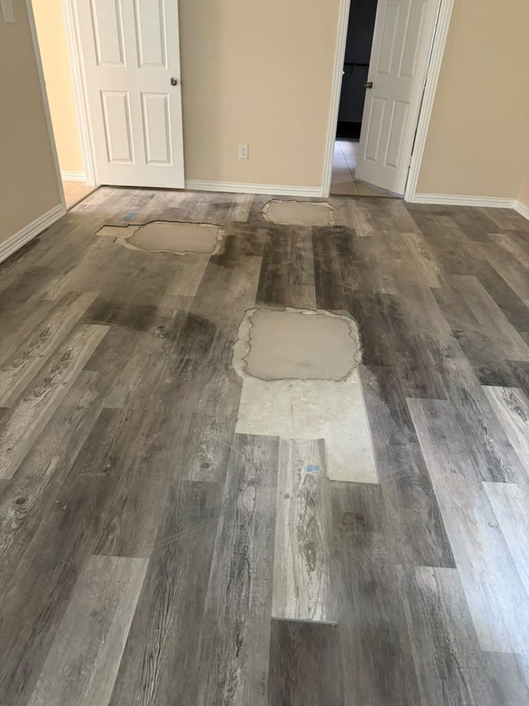 Room with damaged gray wood-look flooring and patched areas near a white door.
