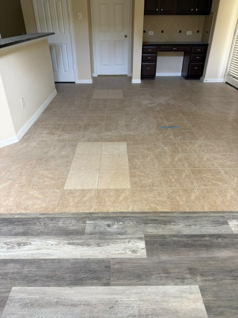 Empty open-plan room with beige tile and wood-look flooring, white doors, and a dark kitchen area in back